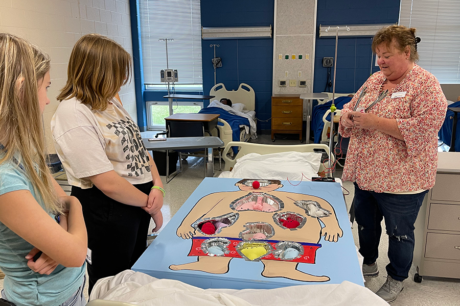 two summer healthcare attendees with a bctc staff member standing over an Operation-style cutout laying in a hospital bed